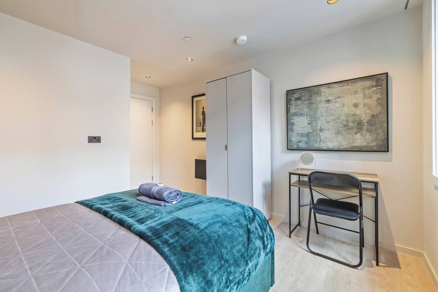 A bedroom with a bed, a desk, a chair, a wardrobe, and a painting on the wall.