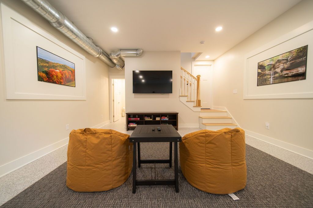 Una habitación con un televisor de pantalla plana montado en la pared, una escalera con barandillas de madera y dos sillones tipo puff.