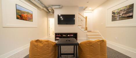 Una habitación con un televisor de pantalla plana montado en la pared, una escalera con barandillas de madera y dos sillones tipo puff.