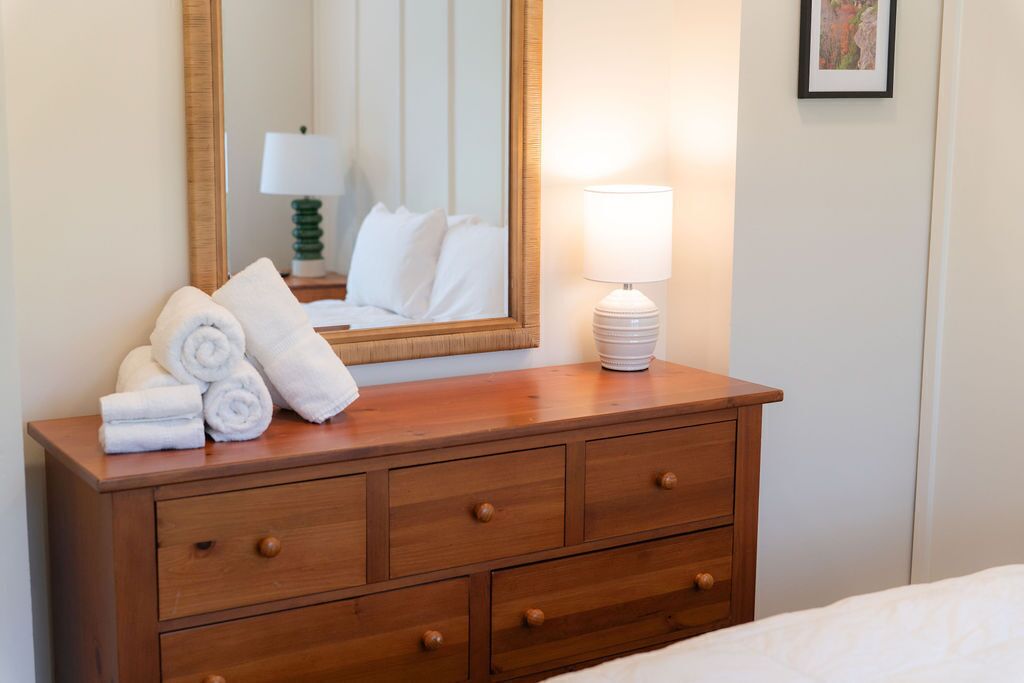 A wooden dresser with rolled towels, a mirror, and a lamp in a hotel room.