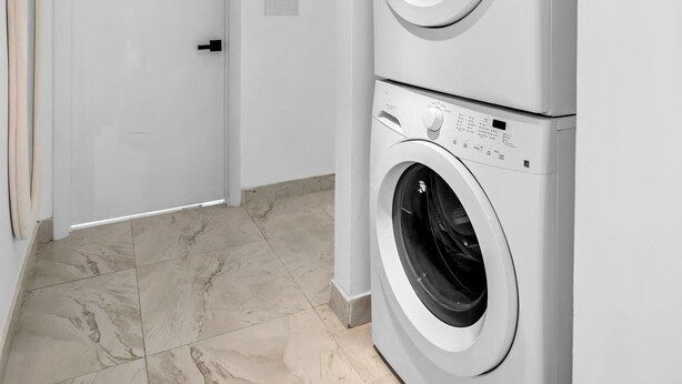 Una lavandería con dos lavadoras de carga frontal apiladas, una puerta blanca y un piso con azulejos.