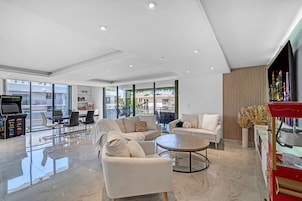 A modern living room with a sofa, armchairs, a coffee table, and a television. There is a balcony with a view of buildings and a pool.
