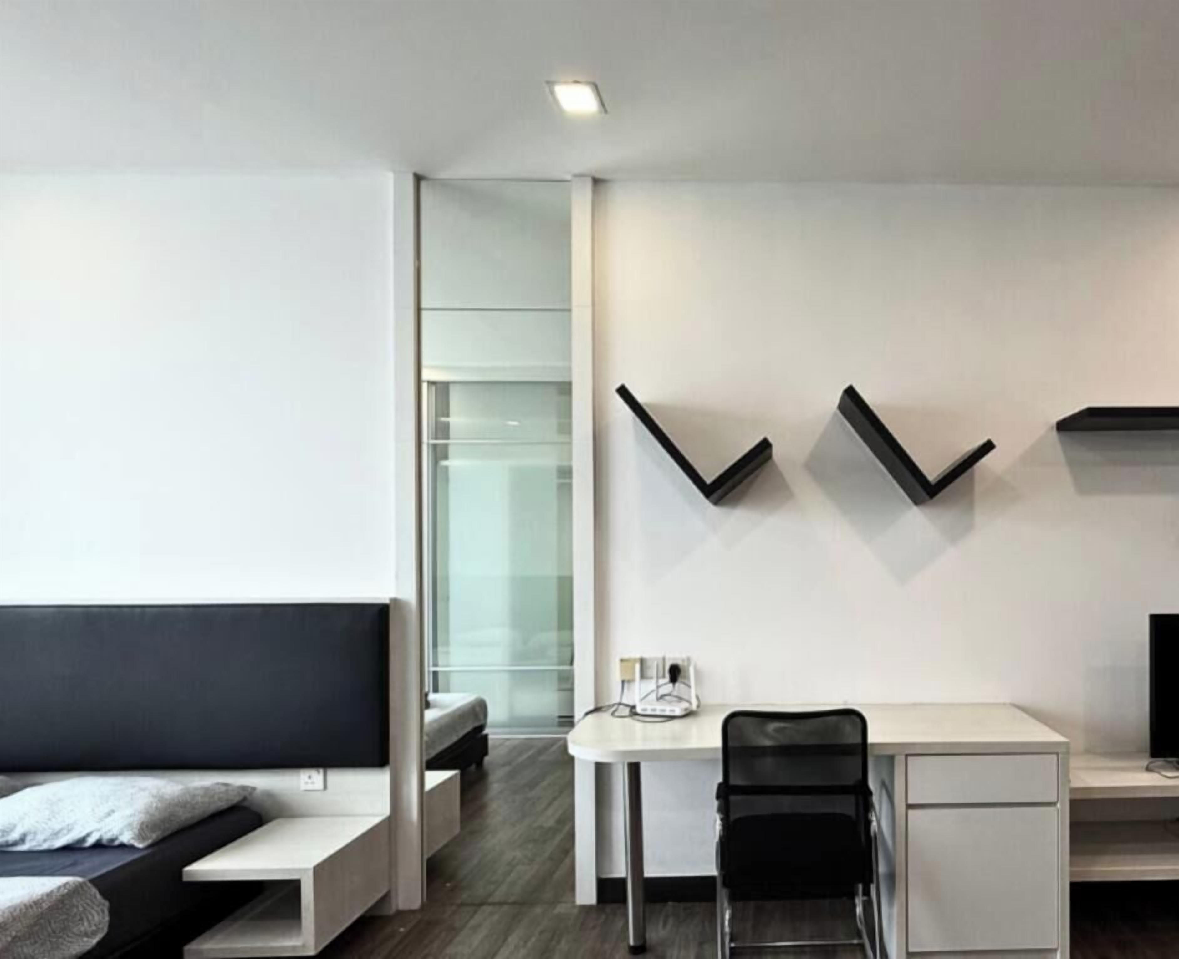 A modern hotel room with a bed, a desk, and a chair. There is a glass partition in the background.