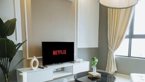 Una sala moderna con un televisor de pantalla plana mostrando 'NETFLIX', un mueble blanco para entretenimiento, una mesa de centro de vidrio, una planta en maceta y un ventanal con cortinas.