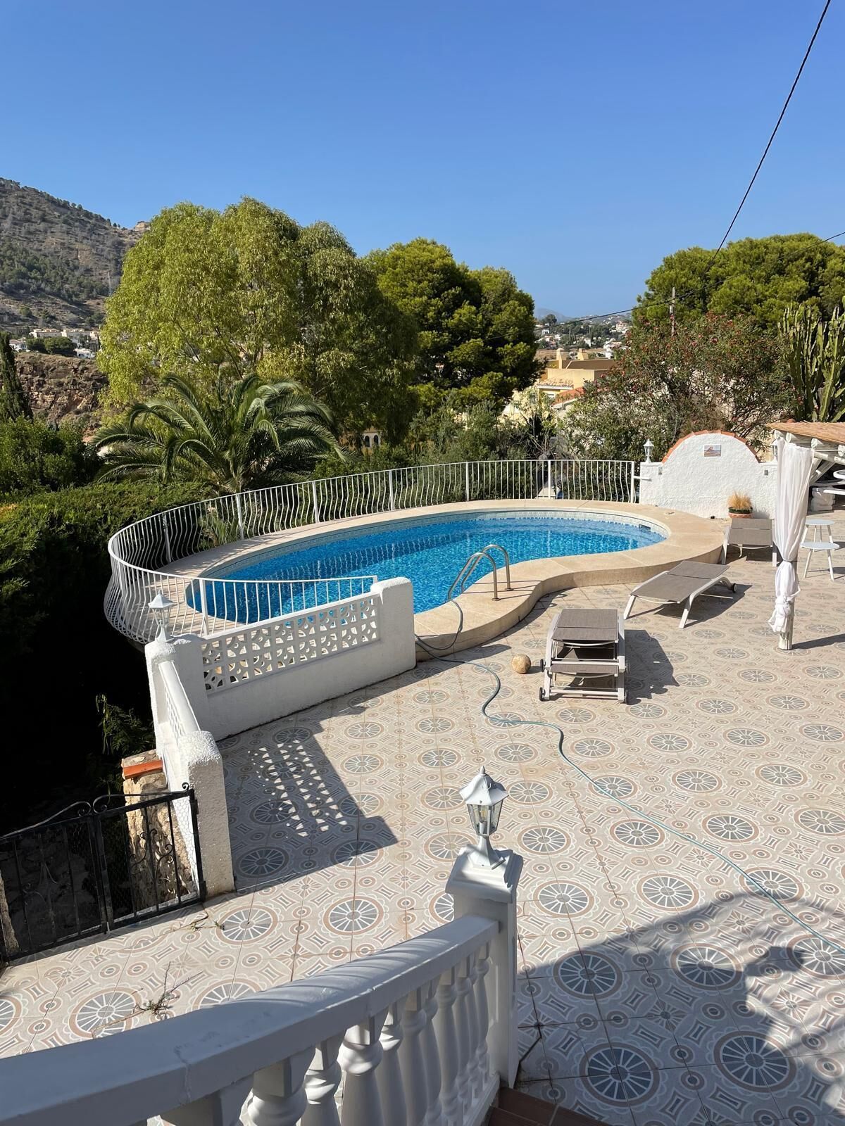 A swimming pool with a white tiled deck, surrounded by a white fence, and overlooking a hilly landscape.
