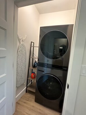A compact laundry room with a front-loading washer and dryer, a drying rack, and a shelf with a bottle of detergent.