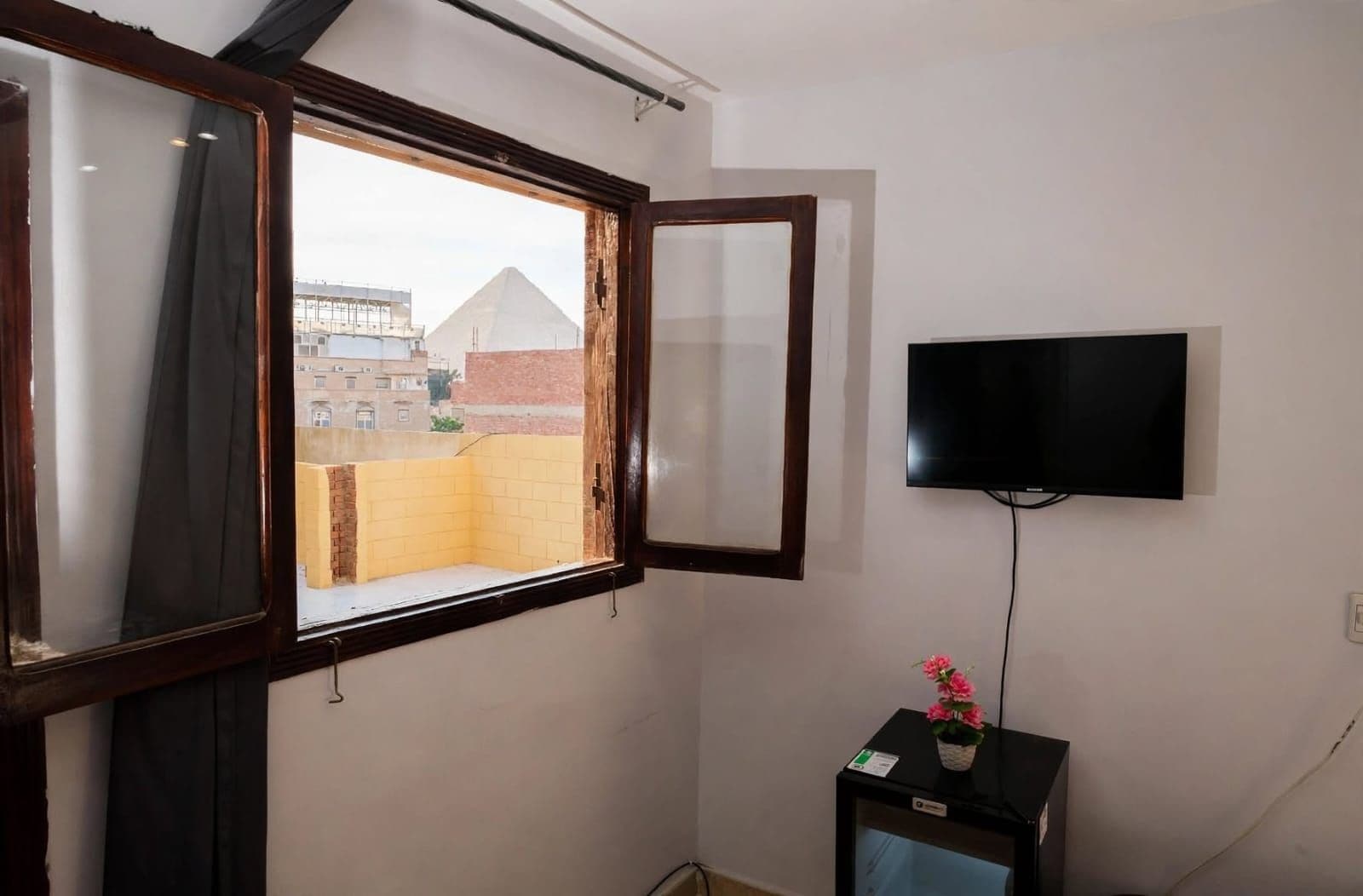 A room with a window, a TV mounted on the wall, and a small table with a flower.