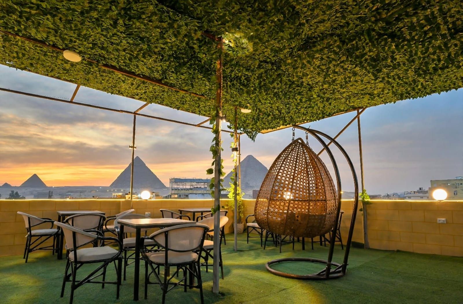 An outdoor seating area with tables and chairs, a hanging chair, and a view of pyramids in the background.