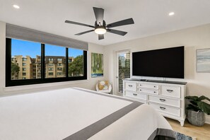 Une chambre avec un grand lit, un ventilateur de plafond, une télévision et une fenêtre donnant sur des immeubles.