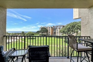 A balcony with wicker furniture, a clear view of a golf course, and a building complex in the background. - Sunset Rentals | Island Club 3302 (Hilton Head Island)