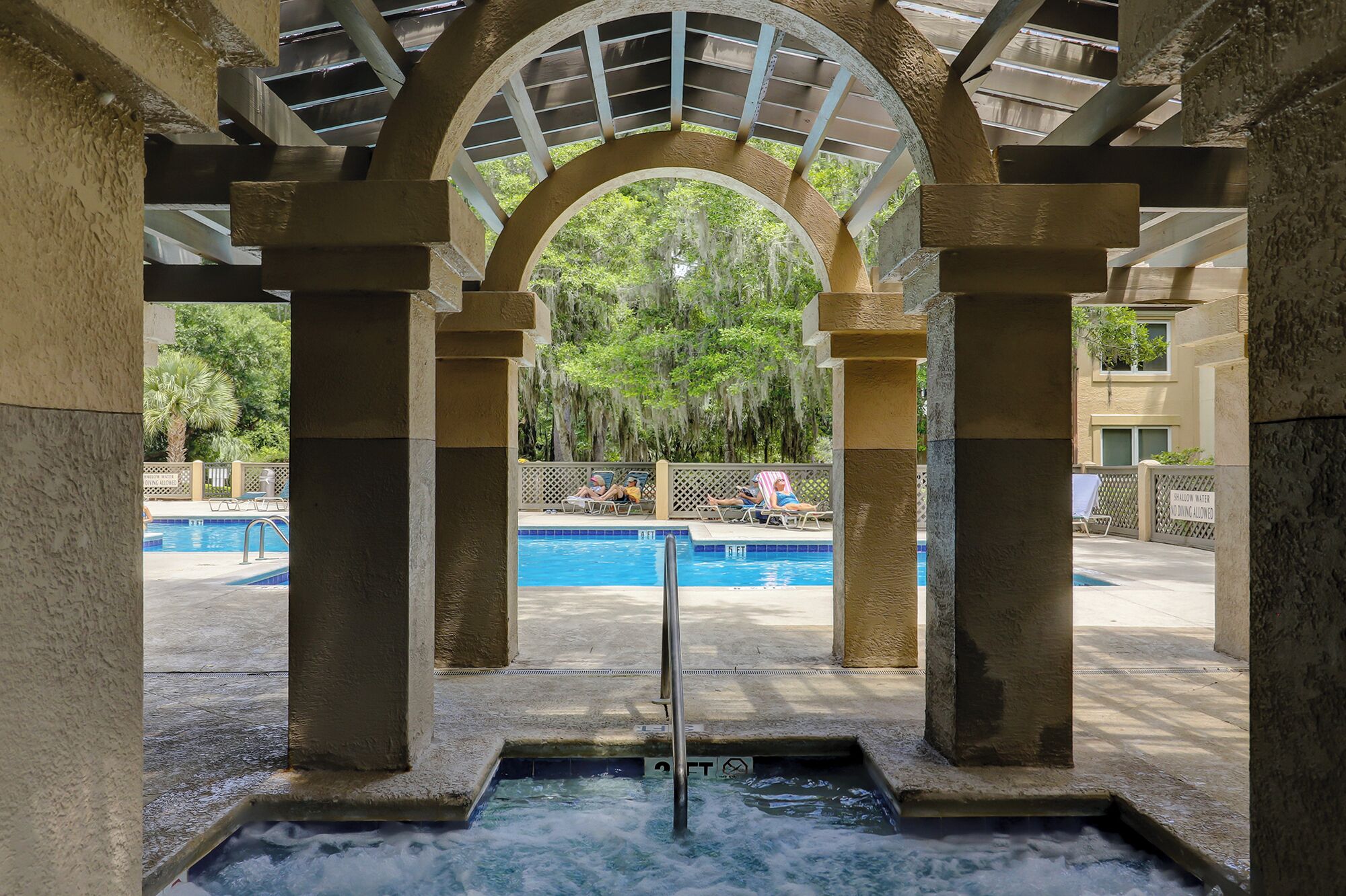Un espace avec un bain à remous, une piscine en arrière-plan, le tout entouré de colonnes et d’une allée couverte.