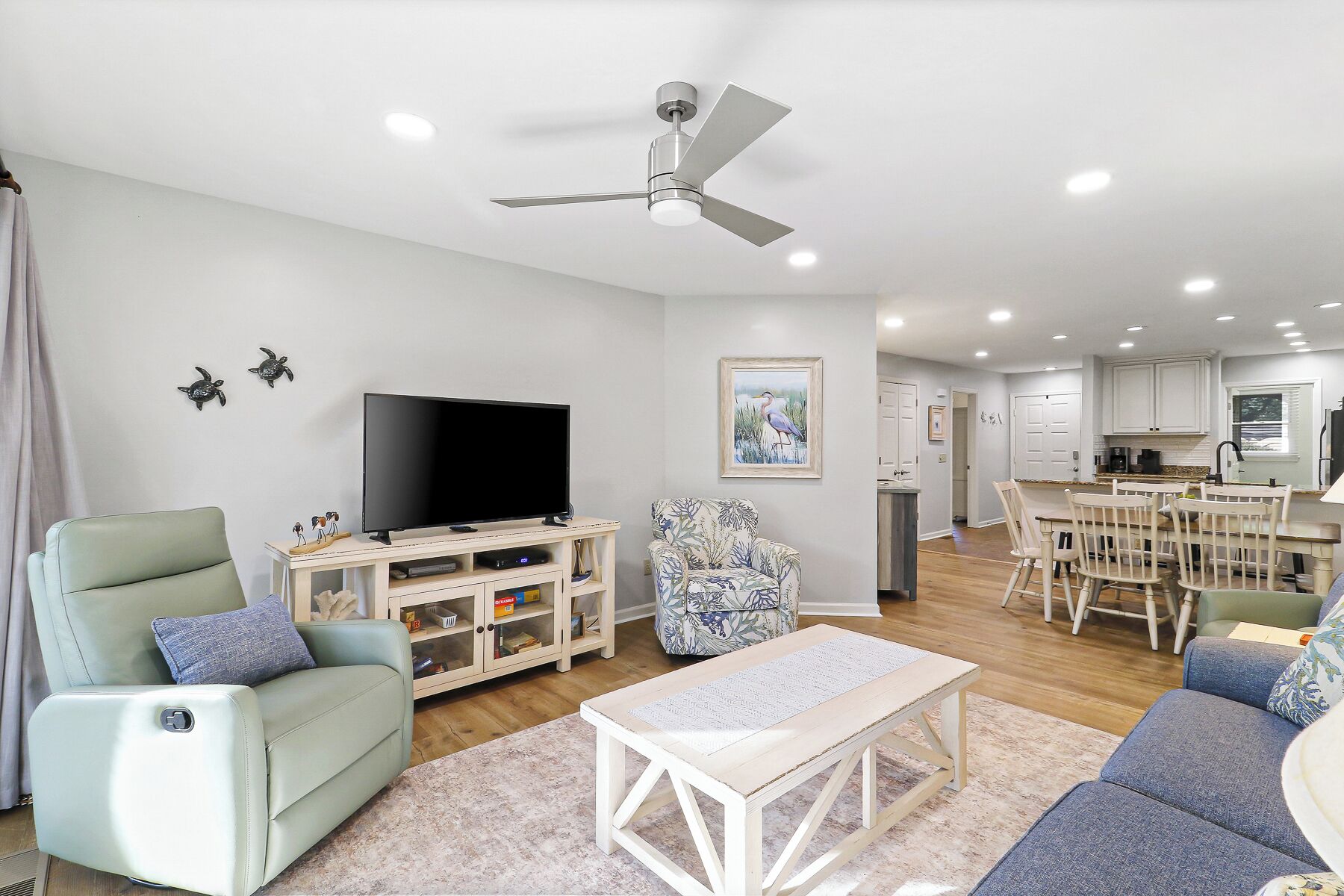 A modern living room with a flat-screen TV, a ceiling fan, a dining area with a table and chairs, and a kitchen in the background.