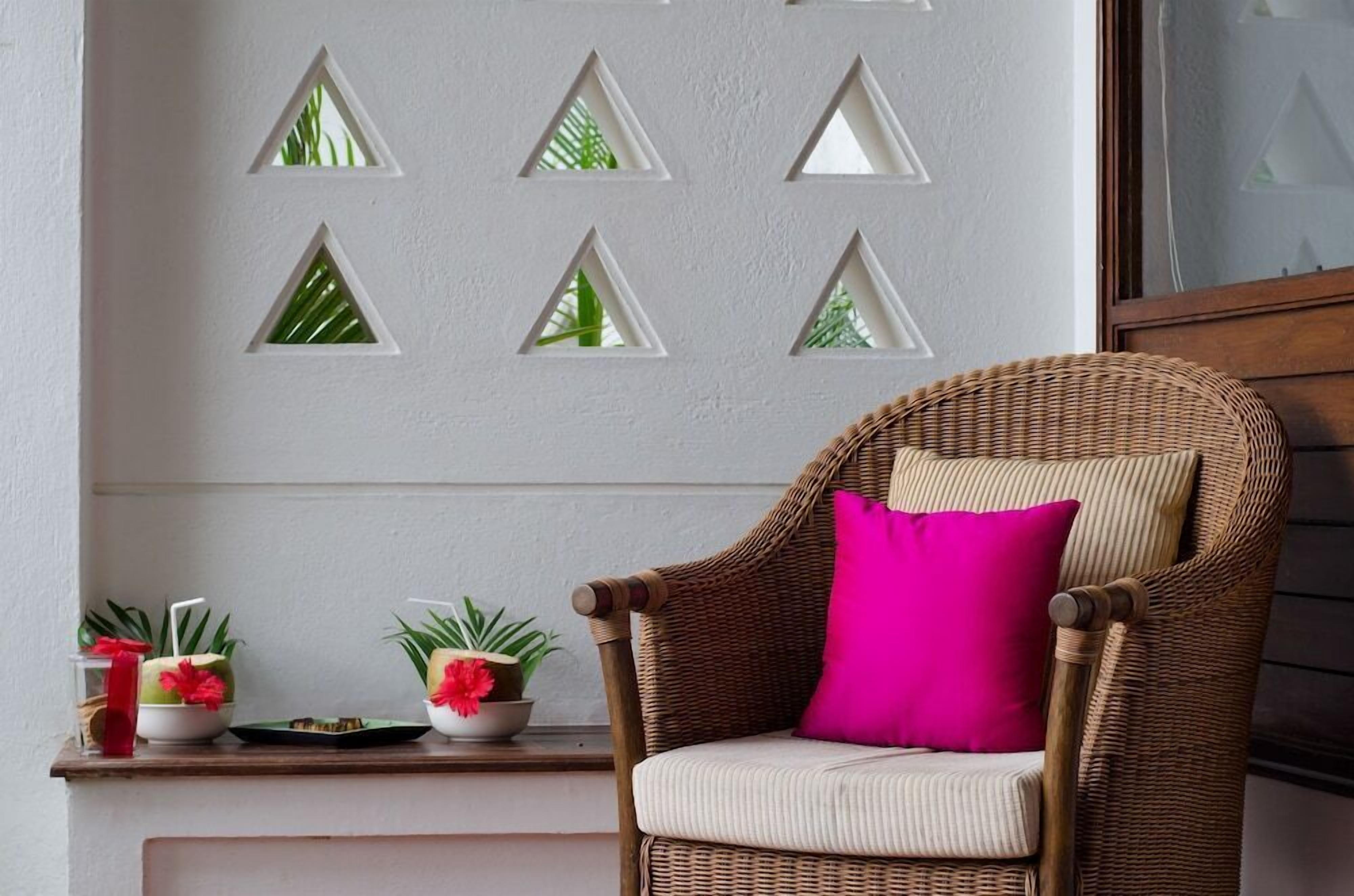 A wicker chair with a cushion and a bright pink pillow, positioned in front of a wall with triangular cutouts.