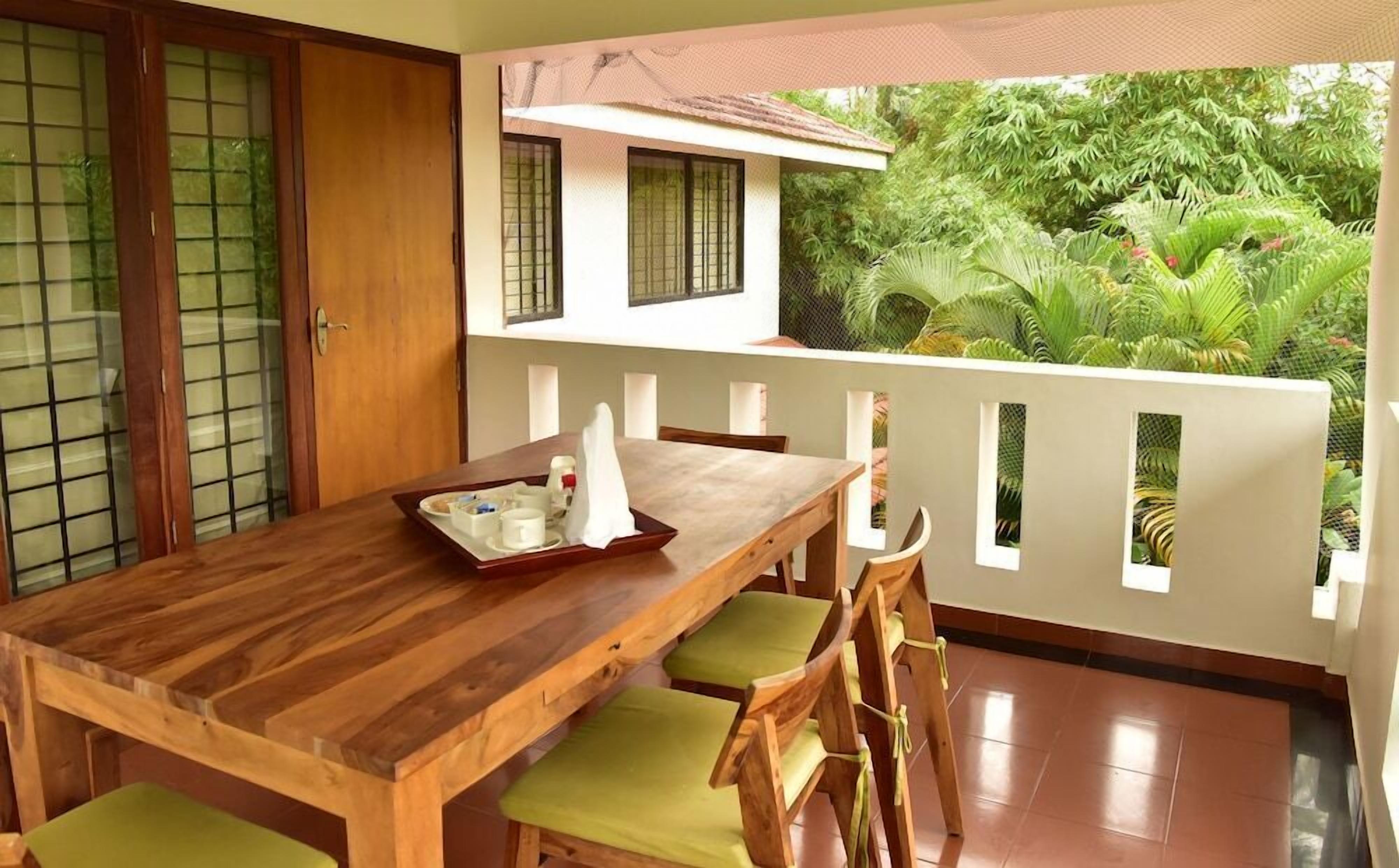 A wooden dining table with chairs, a tray with cups and a saucer, and a view of lush greenery through a balcony.