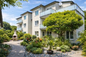 Un edificio residencial de varios pisos con balcones, un camino de entrada pavimentado y una variedad de plantas y arbustos.