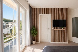 A bedroom with a large window, a potted plant, a TV mounted on a wooden panel wall, and a bed. - Villa Manatee - Wellness Apartment with a Balcony (Sellin)