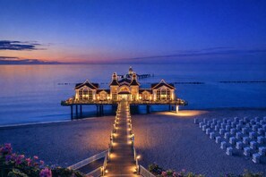 Un hotel frente al mar con un muelle largo que conduce al edificio, una playa con filas de sillas y un mar tranquilo.