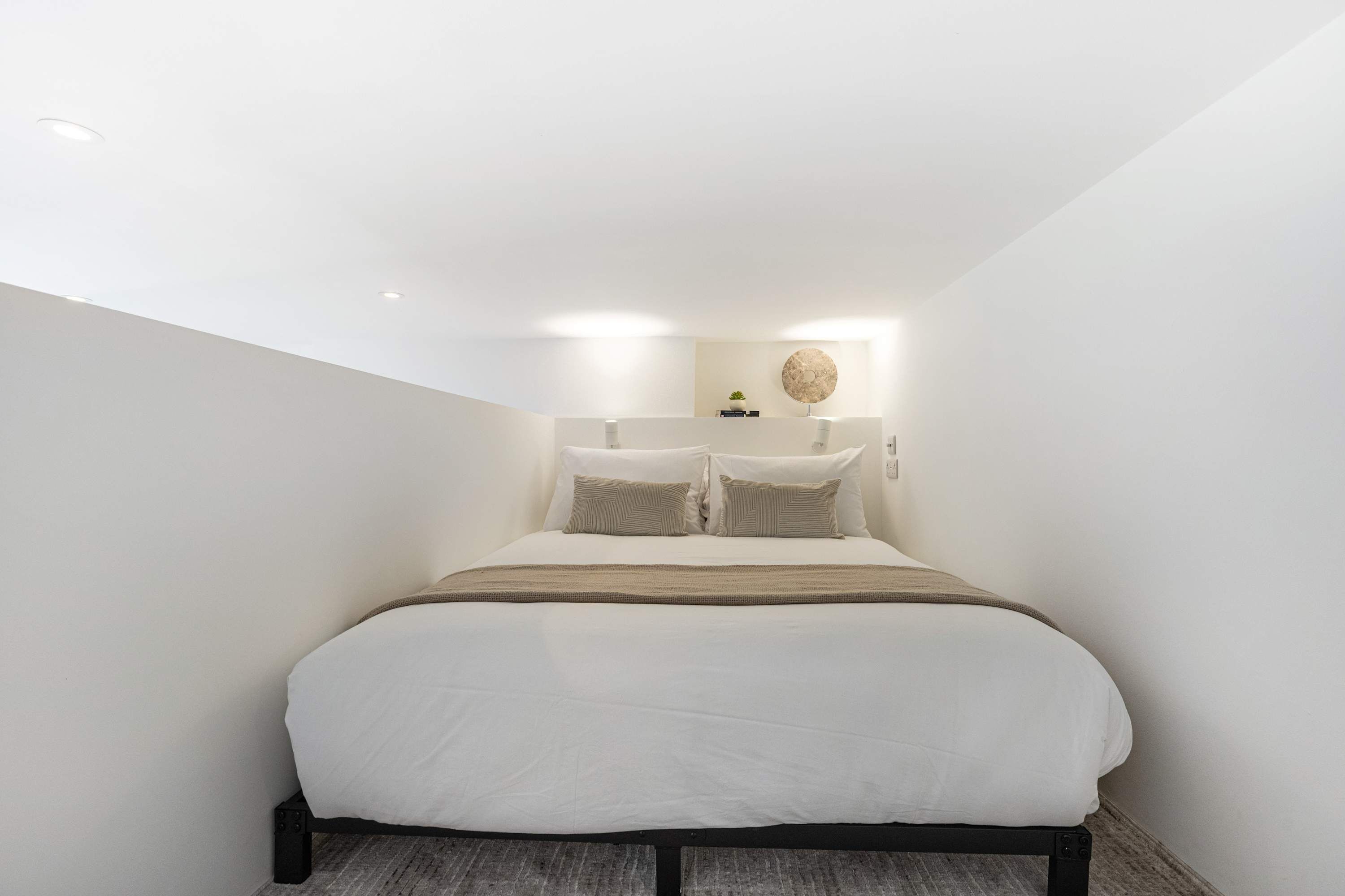 Quarto moderno com uma cama grande, uma mesinha de cabeceira, uma luminária de parede e um pequeno vaso de planta.