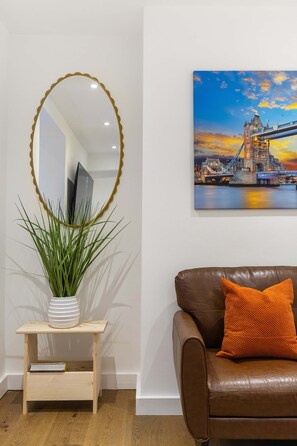 A room with a brown leather sofa, a white vase with green plants, a wooden side table, a large mirror with a gold frame, and a painting of a bridge. - Primrose Hill 1BR Flat - Private Patio (Greater London)