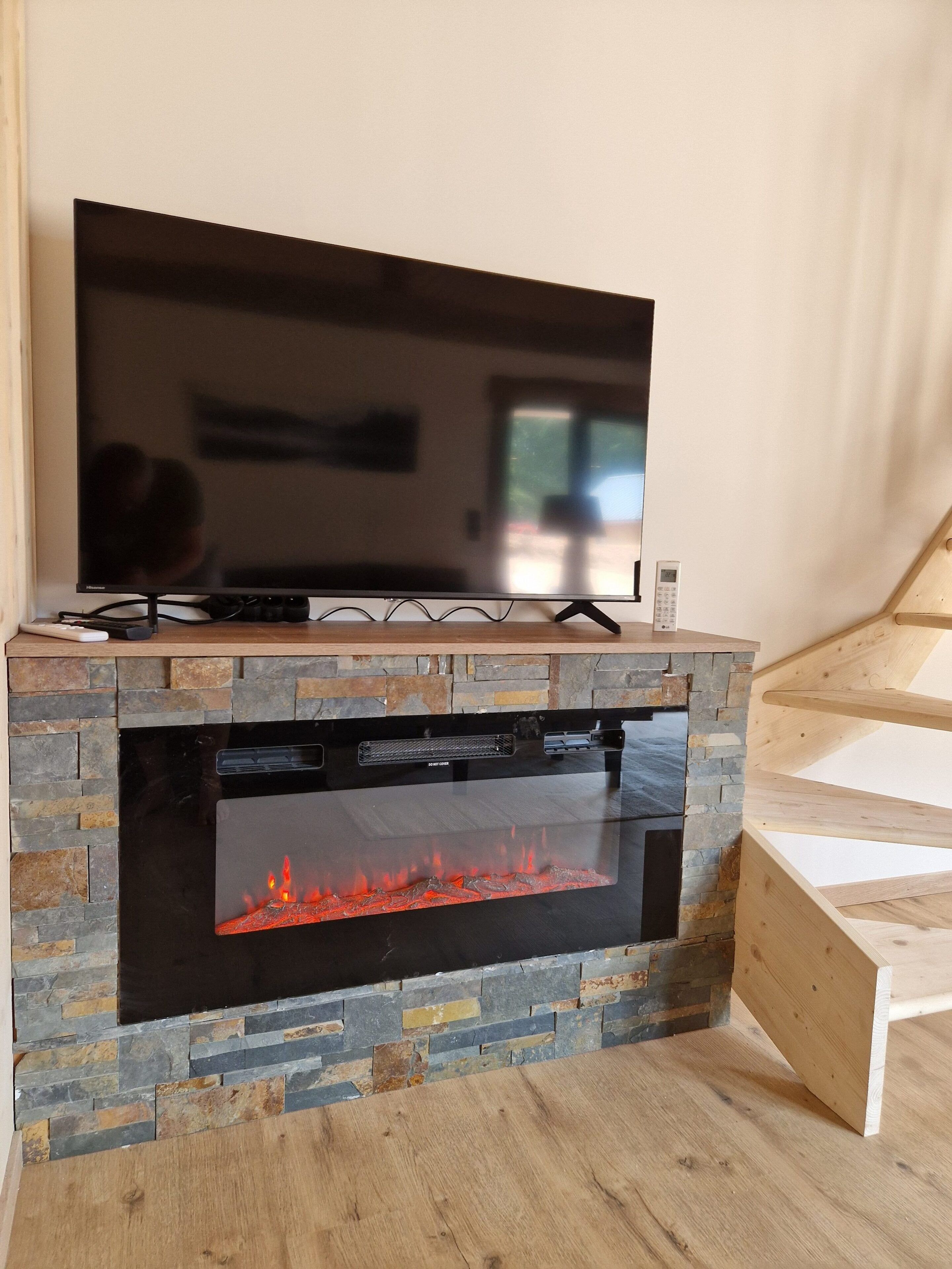 A flat-screen TV mounted on a stone fireplace with a glass door, and a wooden staircase to the right.