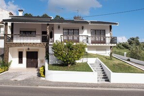 Ein zweistöckiges Haus mit einer Stein-Fassade, einem Hauseingang mit Doppeltüren, einem Balkon im oberen Geschoss und einem kleinen Garten.