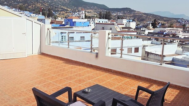 A terrace with two chairs and a table, overlooking a residential area with houses and a hill in the background. - Coffee baba Chefchaouen (Chefchaouen)