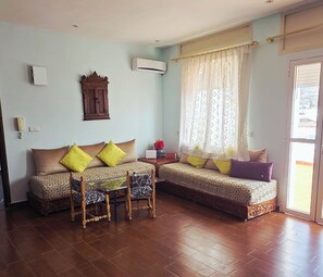 A living room with a sofa set, a small table, and a chair. There is a window with curtains and an air conditioning unit on the wall. - Coffee baba Chefchaouen (Chefchaouen)