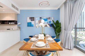 A dining area with a wooden table set for four, blue chairs, and a large window with a view of buildings. - bnbme - Dubai Creek Residences - 2BR (Dubai)