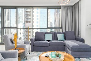 A modern living room with a grey sectional sofa, a small side table, and a large window offering a city view. - bnbme - Dubai Creek Residences - 2BR (Dubai)