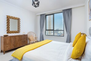 A hotel room with a bed, a wooden dresser, a chair, a window with curtains, and a city view. - bnbme - Dubai Creek Residences - 2BR (Dubai)