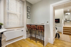 A modern kitchen with a white countertop, three bar stools, and a window with blinds. - Central Russell Square Studio Retreat (Greater London)