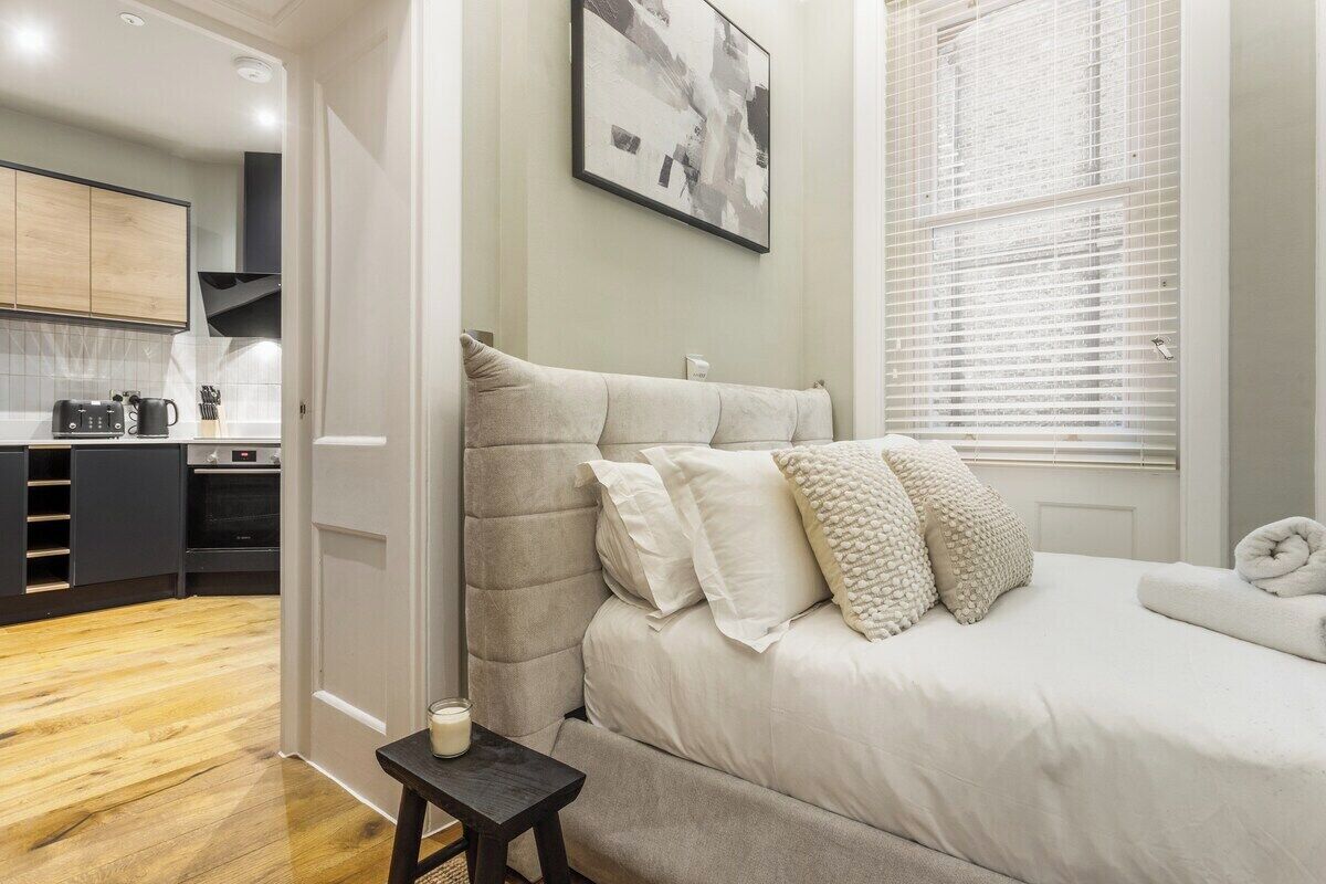 A bedroom with a bed, bedside table, and a view of a kitchen and living area.