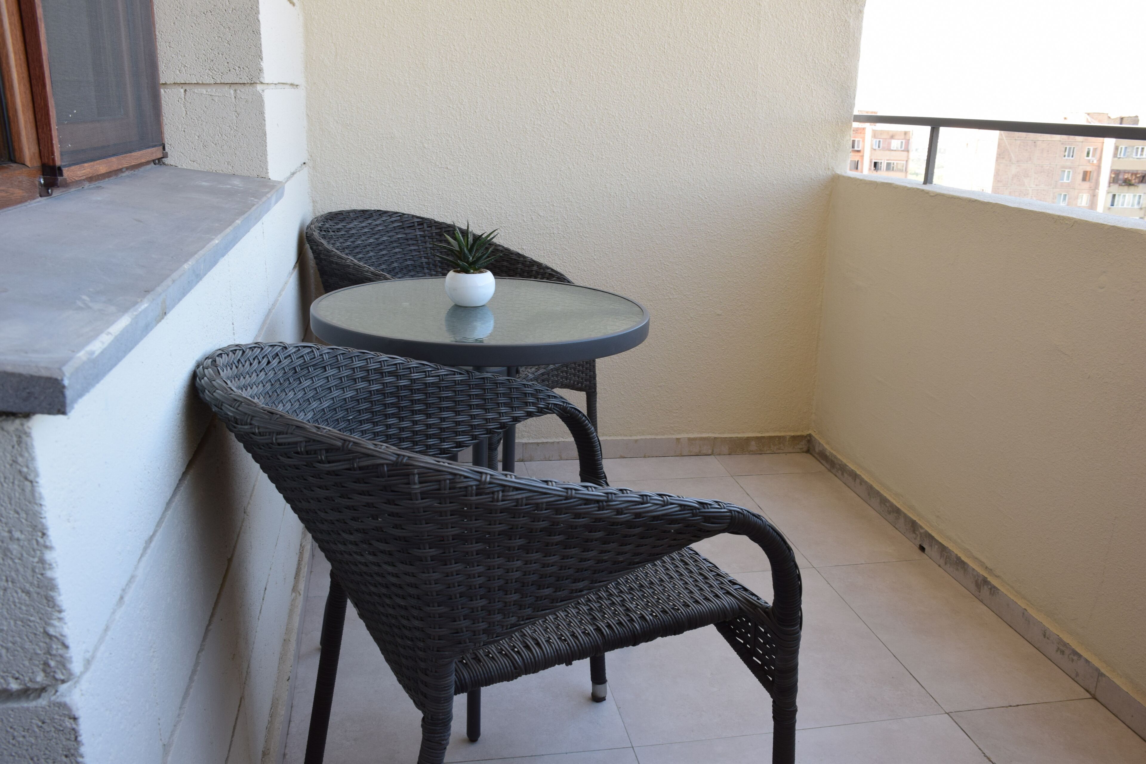 A small balcony with a round table and two wicker chairs, a potted plant on the table, and a window overlooking a building.