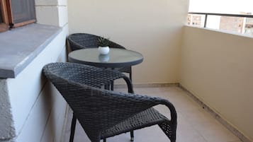 A small balcony with a round table and two wicker chairs, a potted plant on the table, and a window overlooking a building.