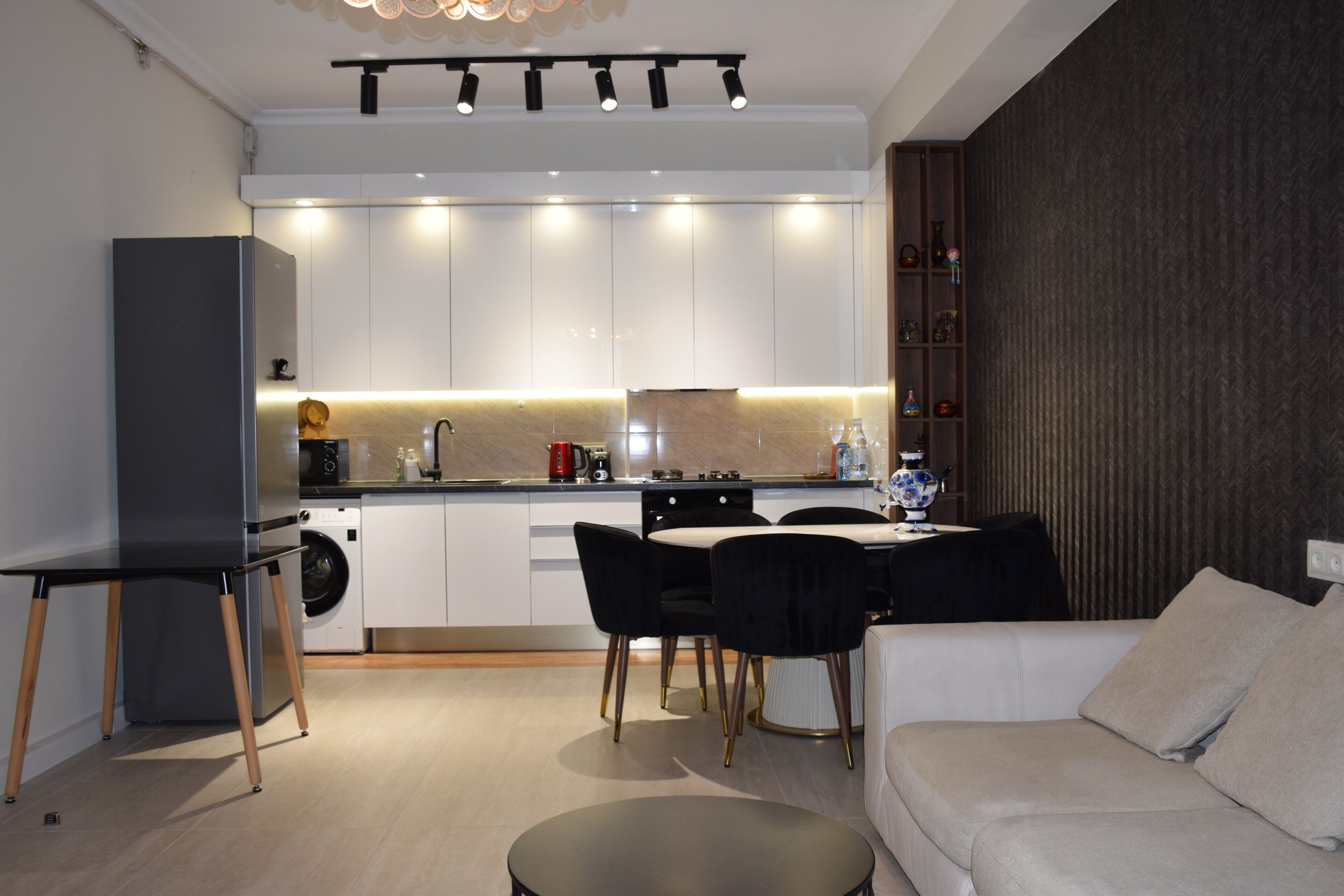 A modern kitchen with white cabinets, a dining area with a black table and chairs, and a sofa with cushions.
