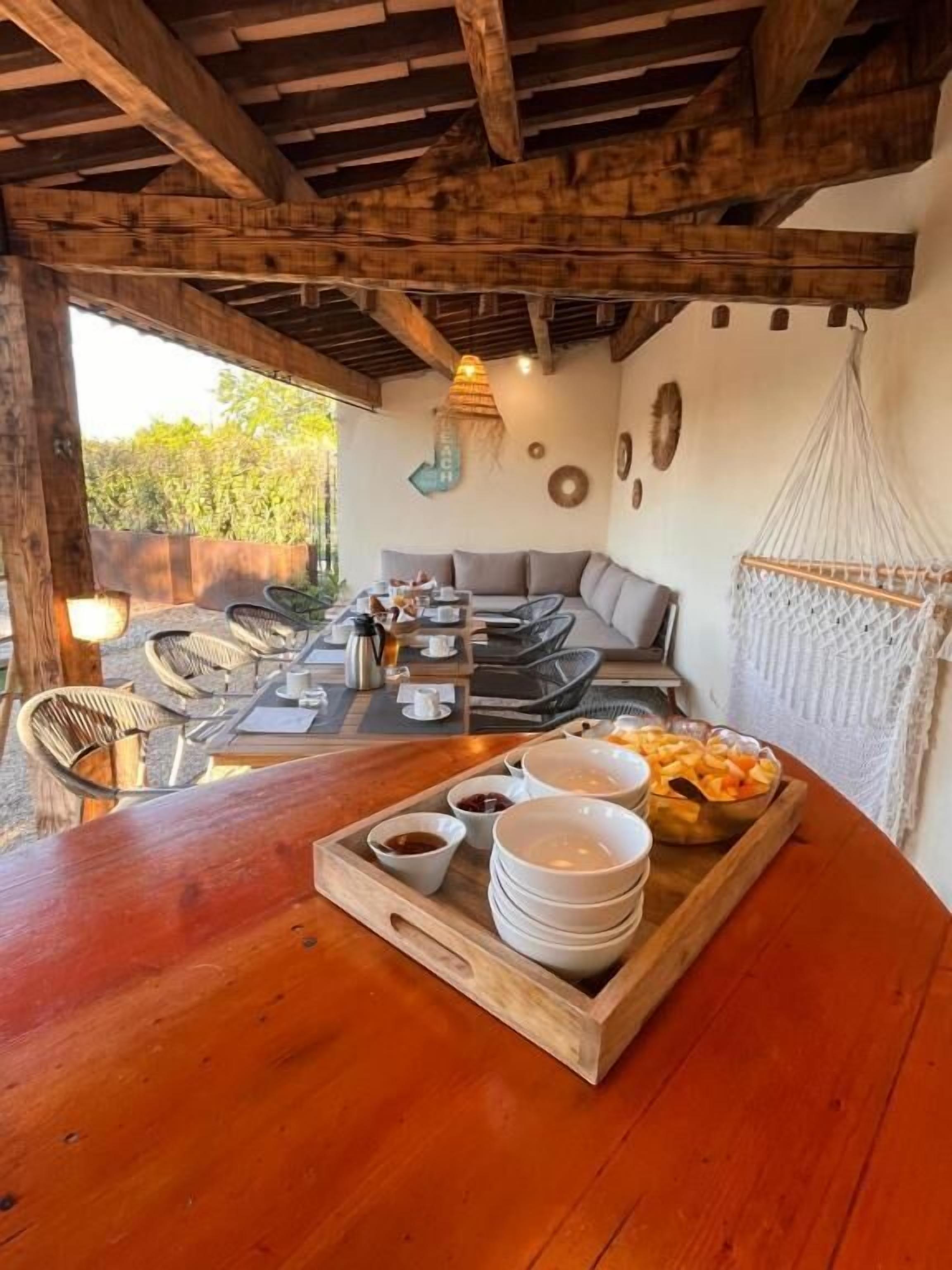 Uma mesa de jantar de madeira com uma bandeja de tigelas e um cesto de pão, num espaço rústico para refeições ao ar livre, com teto de madeira e uma rede.