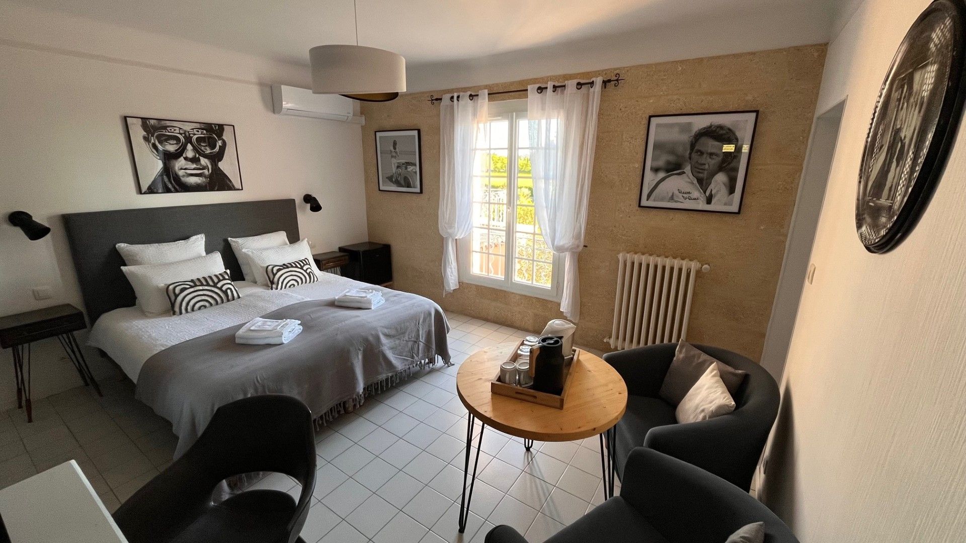 Hotelkamer met een bed, nachtkastjes, een klein rond tafeltje met een koffiezetapparaat, een stoel en een raam met gordijnen.