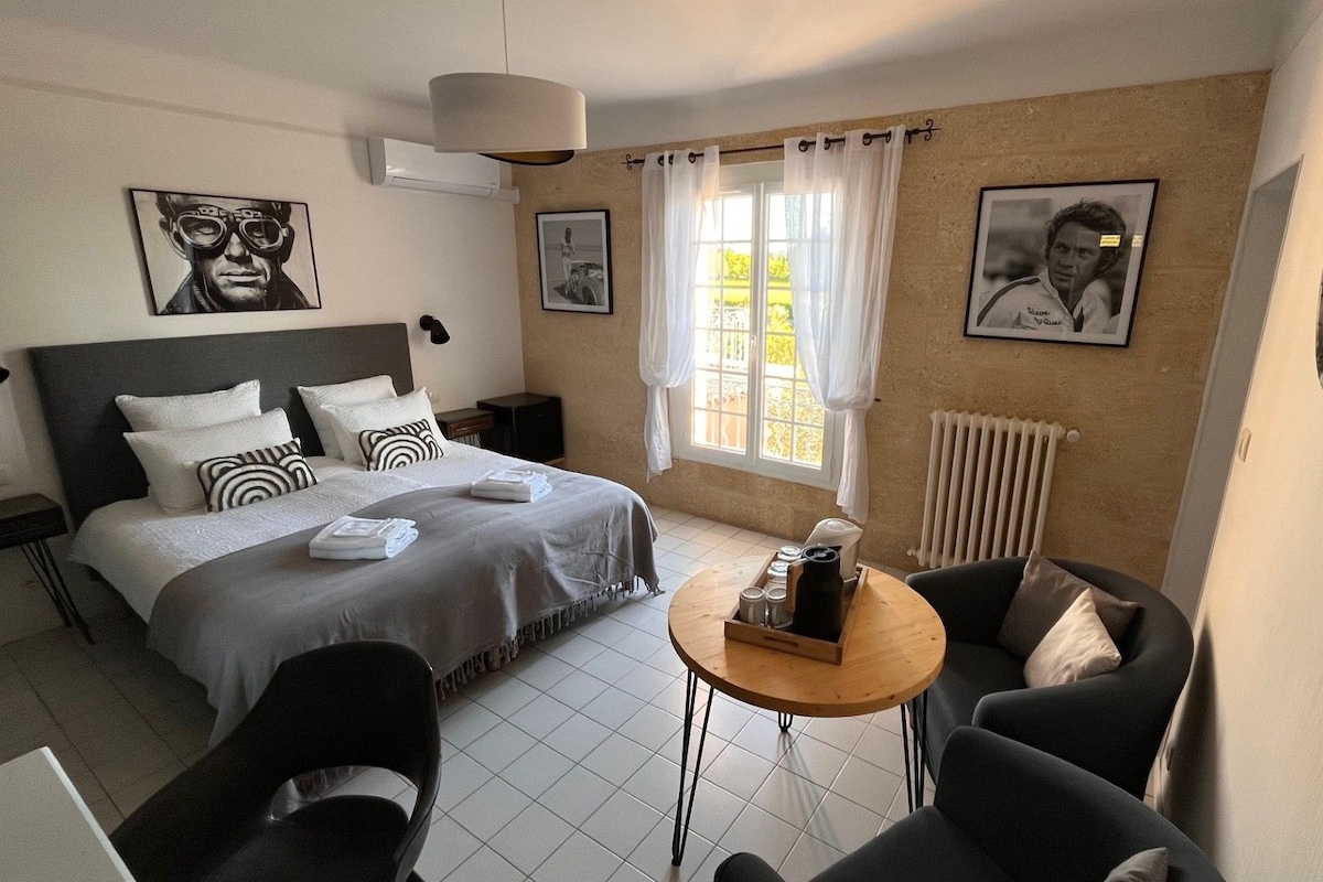 Una camera d'albergo con un letto, un piccolo tavolino rotondo, due poltrone, un termosifone e quadri appesi alle pareti.