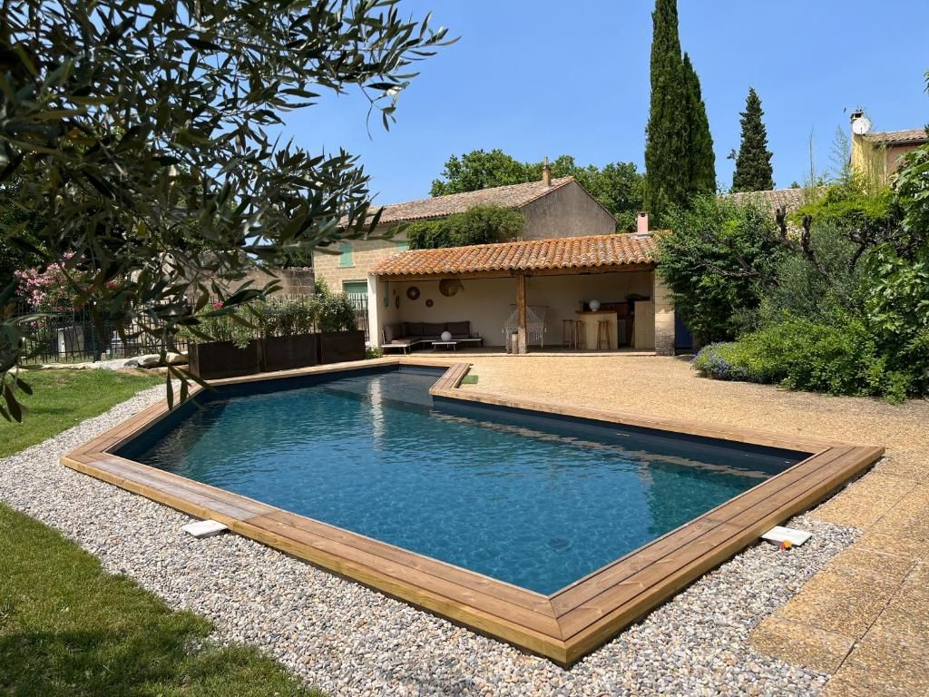 A rectangular swimming pool with a wooden deck, surrounded by gravel and grass, with a house and trees in the background.