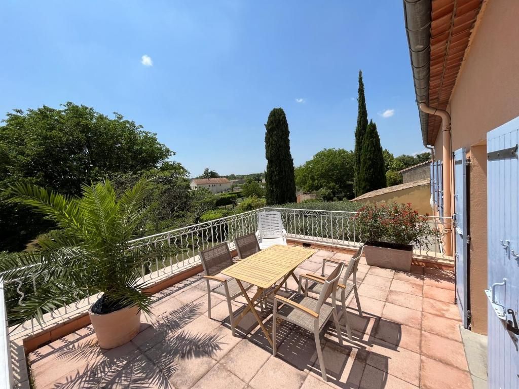 A terrace with a table and chairs, surrounded by a white railing, overlooking a garden with trees and a clear sky.
