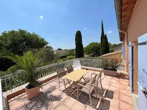 Una terrazza con tavolo e sedie, circondata da un parapetto bianco, affacciata su un giardino con alberi e cielo sereno.