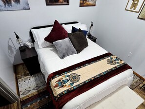 Ein ordentlich bezogenes Bett mit einem gemusterten Überwurf, ein Nachttisch mit Lampe und eine Wand mit gerahmten Bildern.