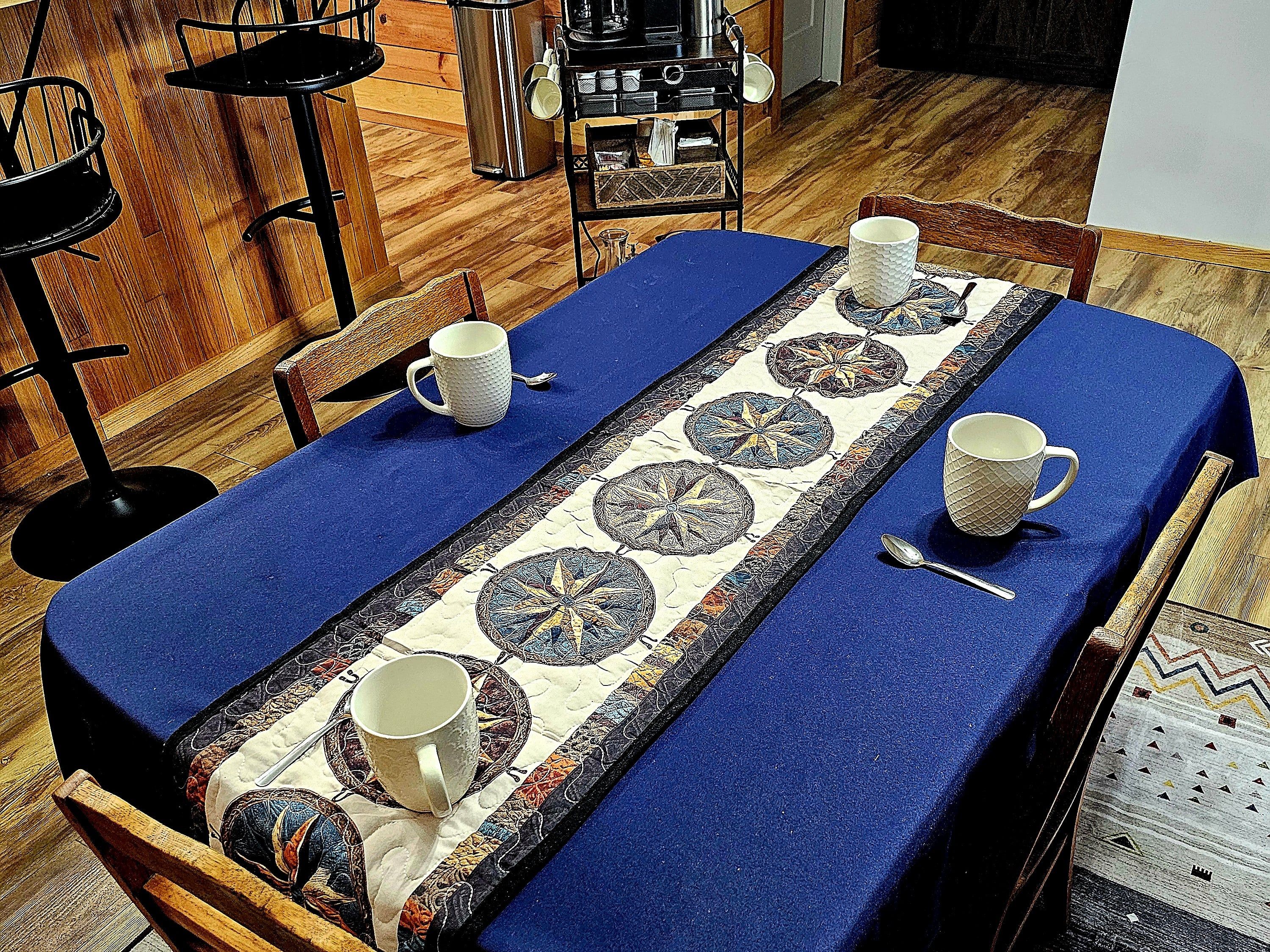 A dining table with a blue tablecloth, a decorative runner, and four white mugs.