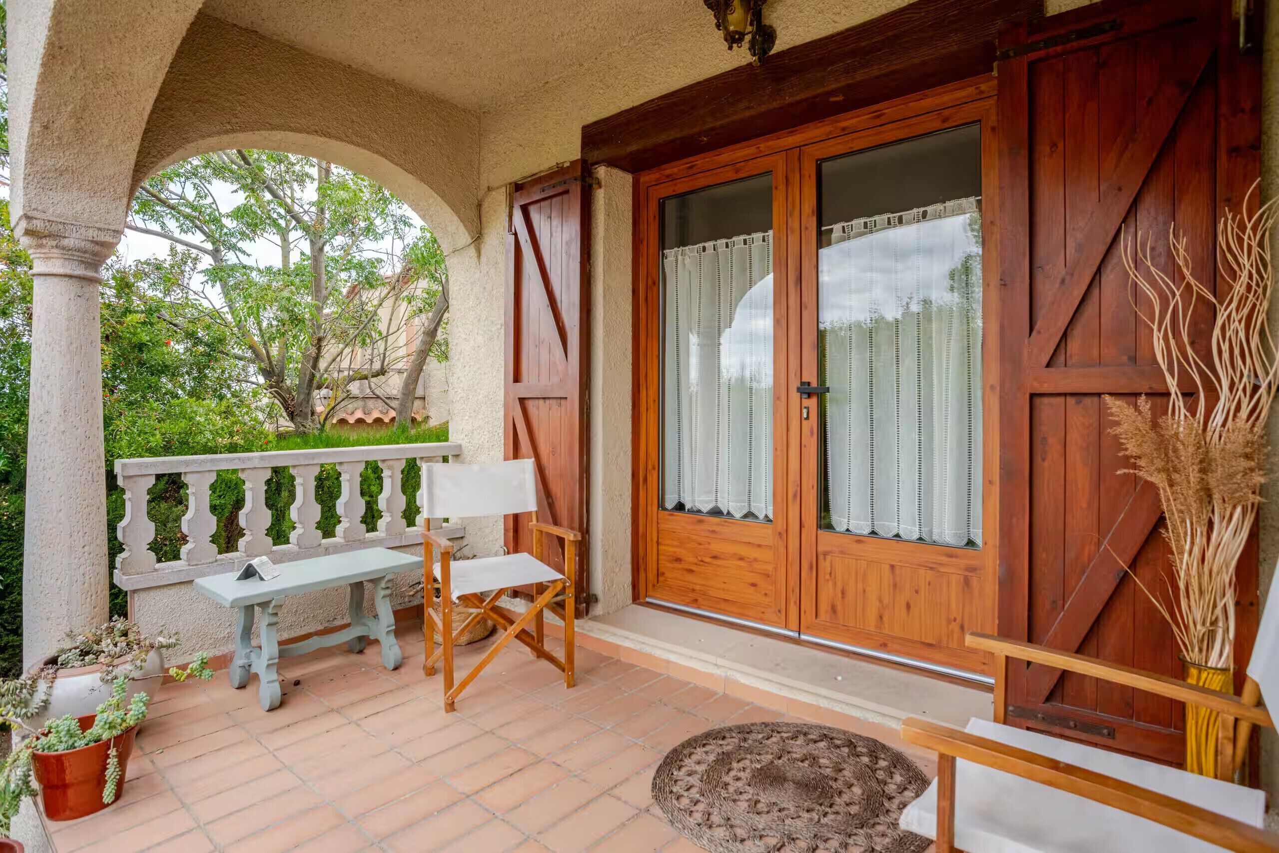 A balcony with a table and chairs, a wooden door with curtains, and potted plants.