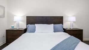 A neatly made bed with a white comforter and two pillows, flanked by two bedside tables with lamps, in a simple room with a plain wall and a wooden headboard. - Villa Bella Monte 1 Bedroom Condo (Phoenix)