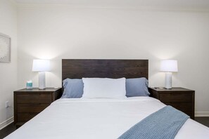 A neatly made bed with a white comforter and two pillows, flanked by two bedside tables with lamps, in a simple room with a plain wall and a wooden headboard. - Villa Bella Monte 1 Bedroom Condo (Phoenix)