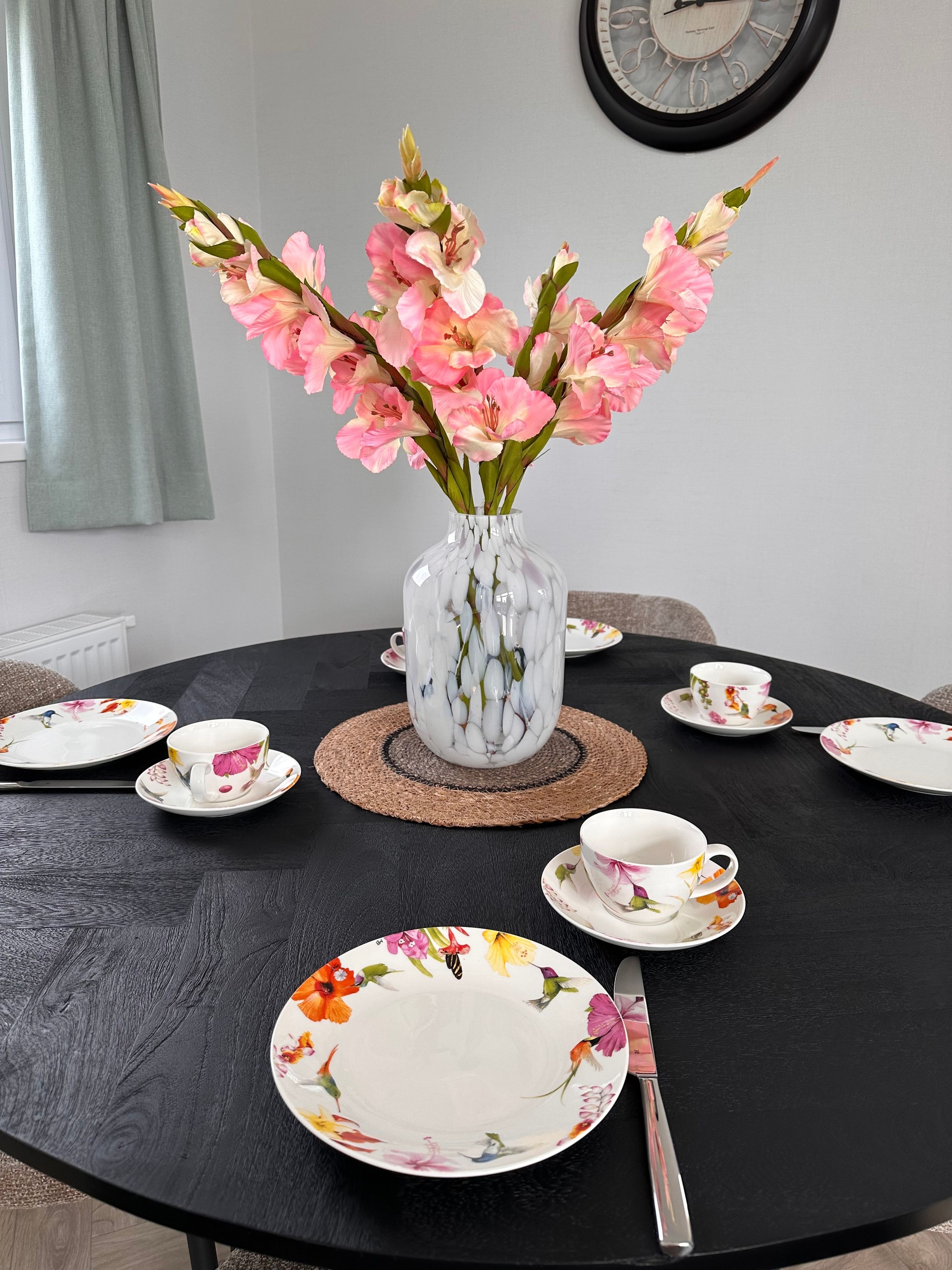 Een ronde eettafel met borden met bloemenprint, een vaas met roze bloemen en een klok aan de muur.