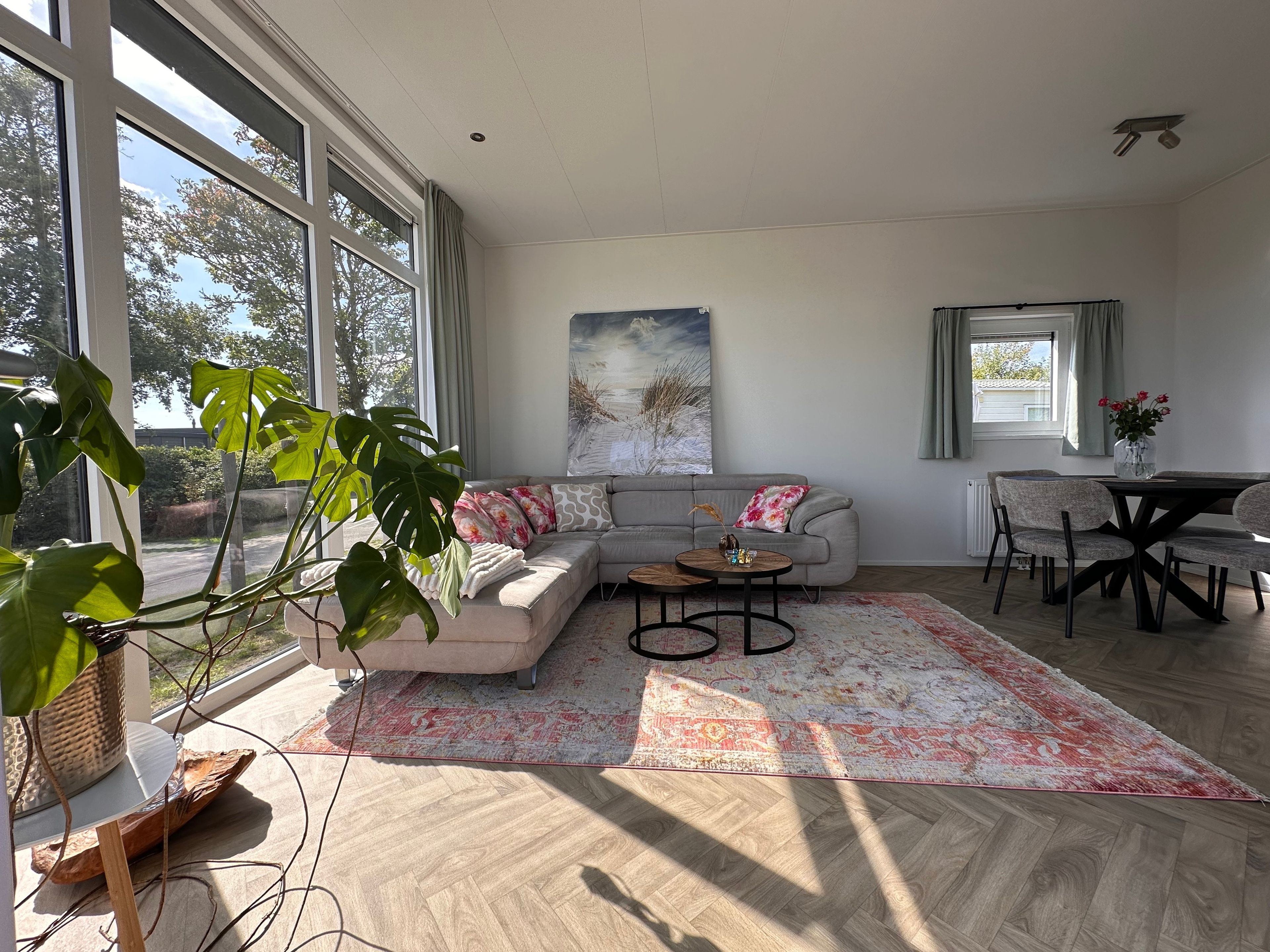 Ein modernes Wohnzimmer mit großem Fenster, einem grauen Sofa, einem Couchtisch und einem Essbereich mit Stühlen.