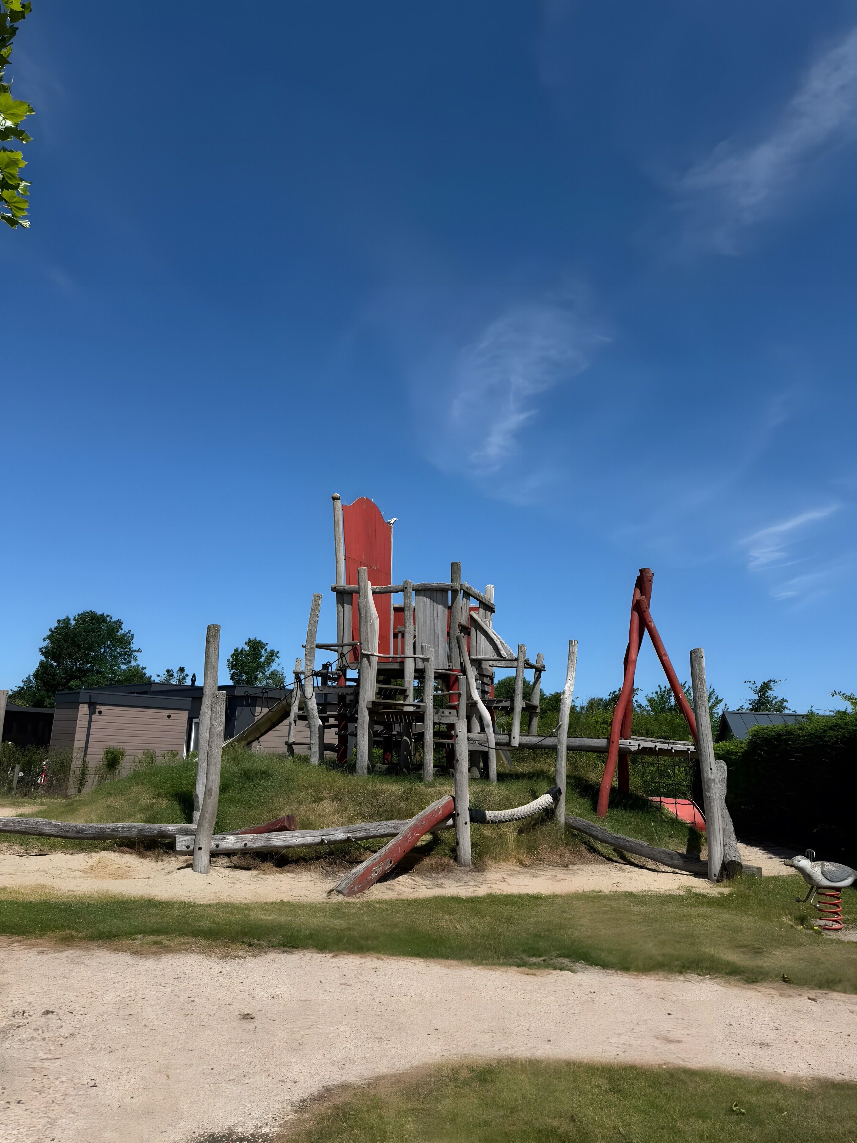 A playground with a slide and climbing structures, surrounded by a grassy area and a dirt path.