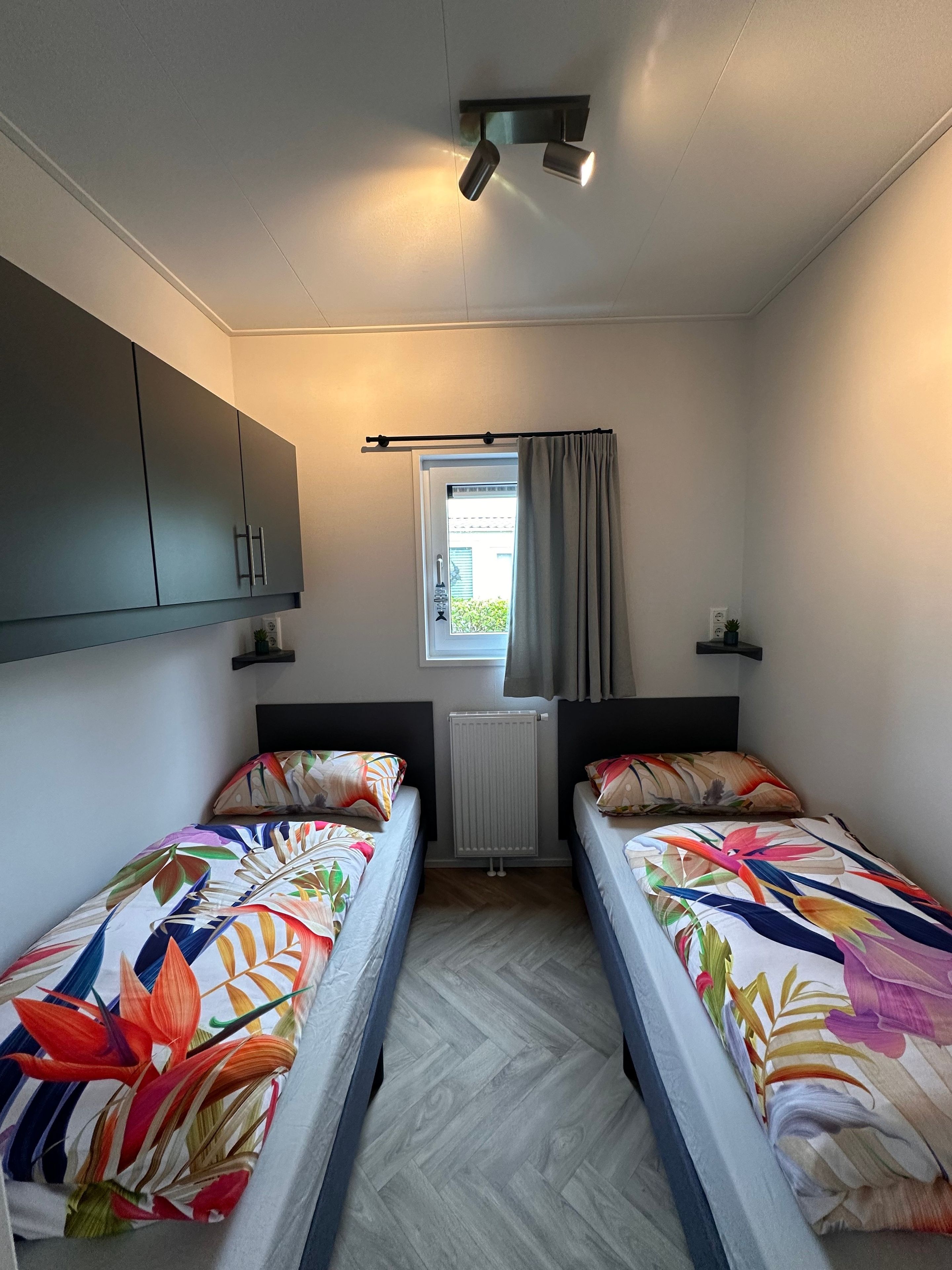 Une chambre avec deux lits, une fenêtre avec des rideaux, un radiateur et un luminaire au plafond.